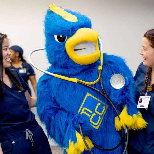 Two nurses in blue scrubs laugh with a large blue and yellow bird mascot wearing a giant stethoscope. The mascot features a "GRCC" logo on its chest.